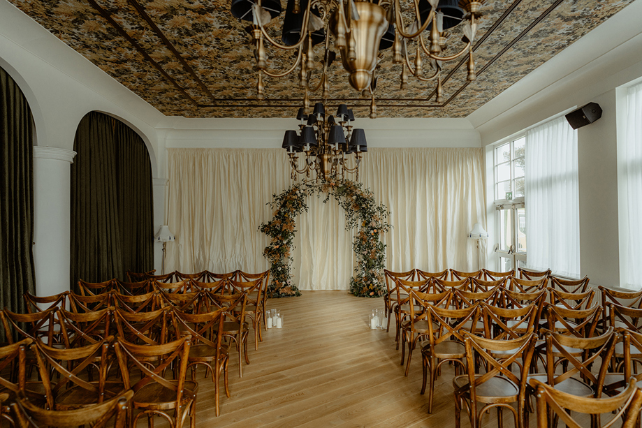 Indoor wedding ceremony setup at Dornoch Station Hotel with wooden cross-back chairs, floral arch and chandelier ceiling