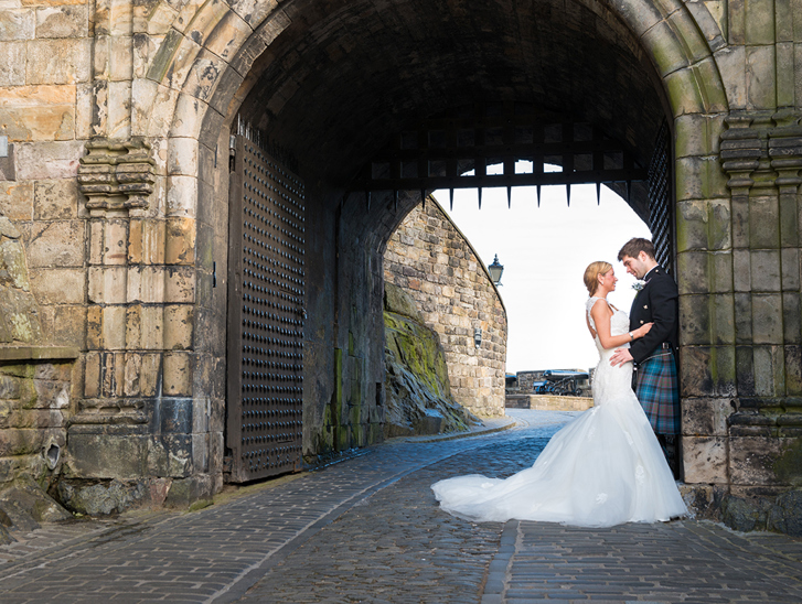Bride and groom standing beneath the historic stone archway entrance at Edinburgh Castle during a romantic Scottish castle wedding photoshoot.