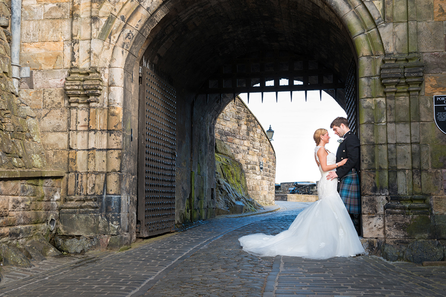 Bride and groom standing beneath the historic stone archway entrance at Edinburgh Castle during a romantic Scottish castle wedding photoshoot.