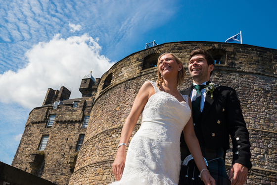 Bride and groom smiling beneath the historic stone walls of Edinburgh Castle with blue skies during their Scottish castle wedding.