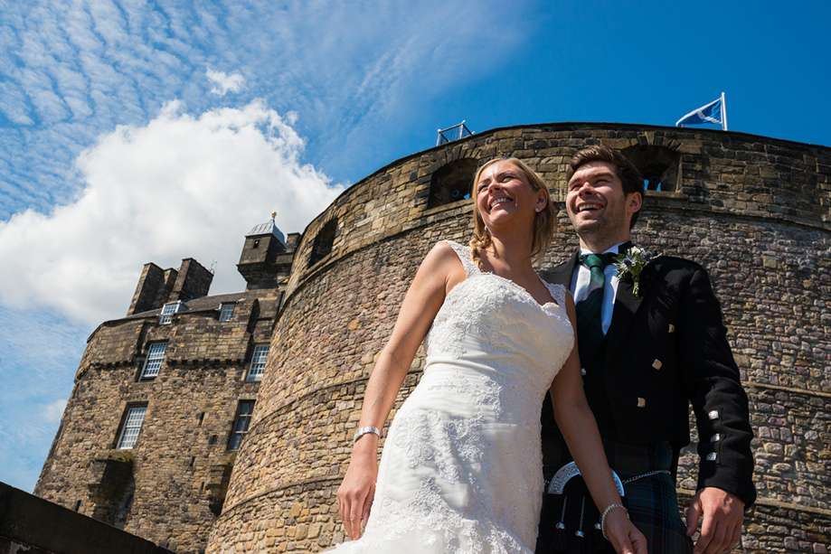 Bride and groom smiling beneath the historic stone walls of Edinburgh Castle with blue skies during their Scottish castle wedding.
