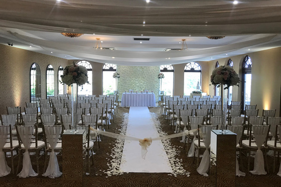 Ceremony room set up with aisle lined with petals, entrance to aisle tied with organza and two bouquets on stands at bottom of aisle