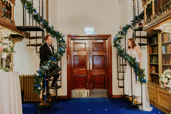 Bride and groom smile at each other from opposite sides of the library spiral staircases, with tinsel and baubles wrapped round bannister
