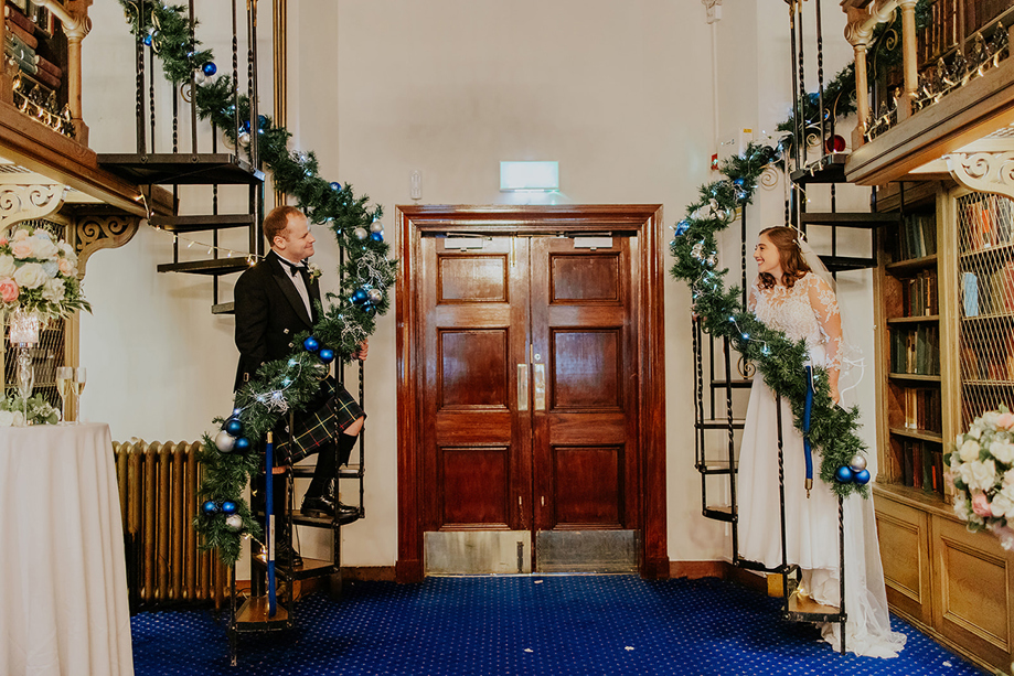 Bride and groom smile at each other from opposite sides of the library spiral staircases, with tinsel and baubles wrapped round bannister