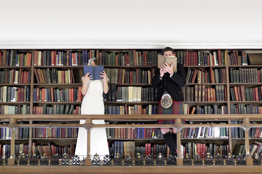 Bride and groom cover their faces with books whilst standing on the upper library