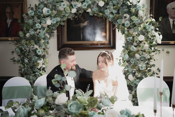 Bride and groom sit at top table in front of floral hoop.