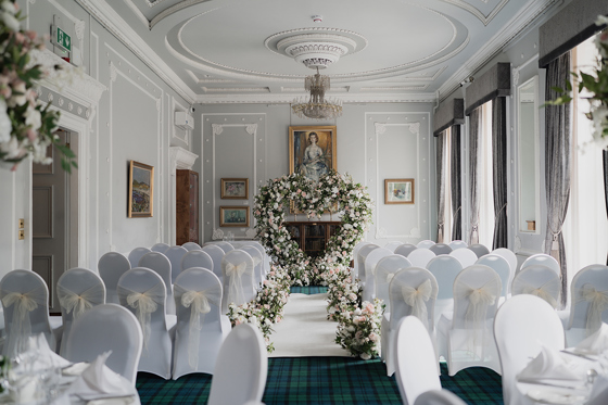 Ceremony set up for photoshoot at 1599 at the Royal College in Glasgow, with white chair covers and cream chiffon bows, and pink and white flowers lining the aisle to a large love heart shaped floral arch at the altar.