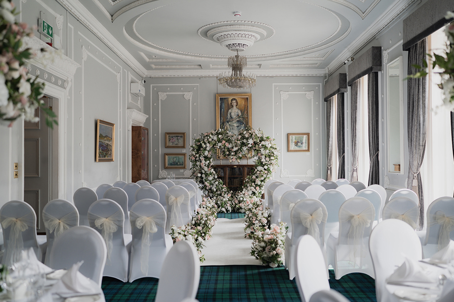 Ceremony set up for photoshoot at 1599 at the Royal College in Glasgow, with white chair covers and cream chiffon bows, and pink and white flowers lining the aisle to a large love heart shaped floral arch at the altar.