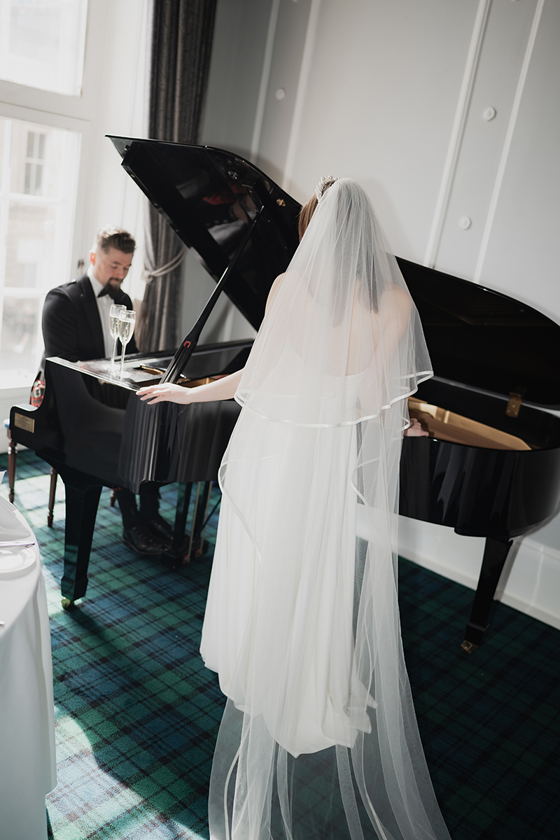 Groom plays grand piano while bride watches on.