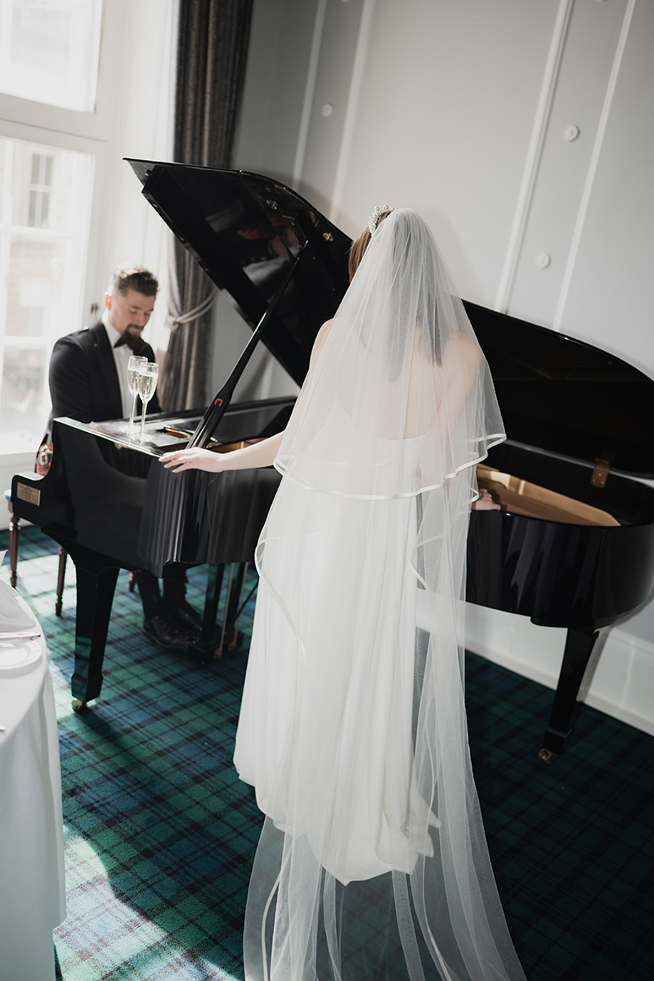 Groom plays grand piano while bride watches on.