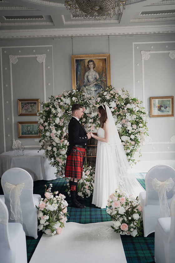 Bride and groom at altar, holding hands in front of a love heart shaped flower arch.
