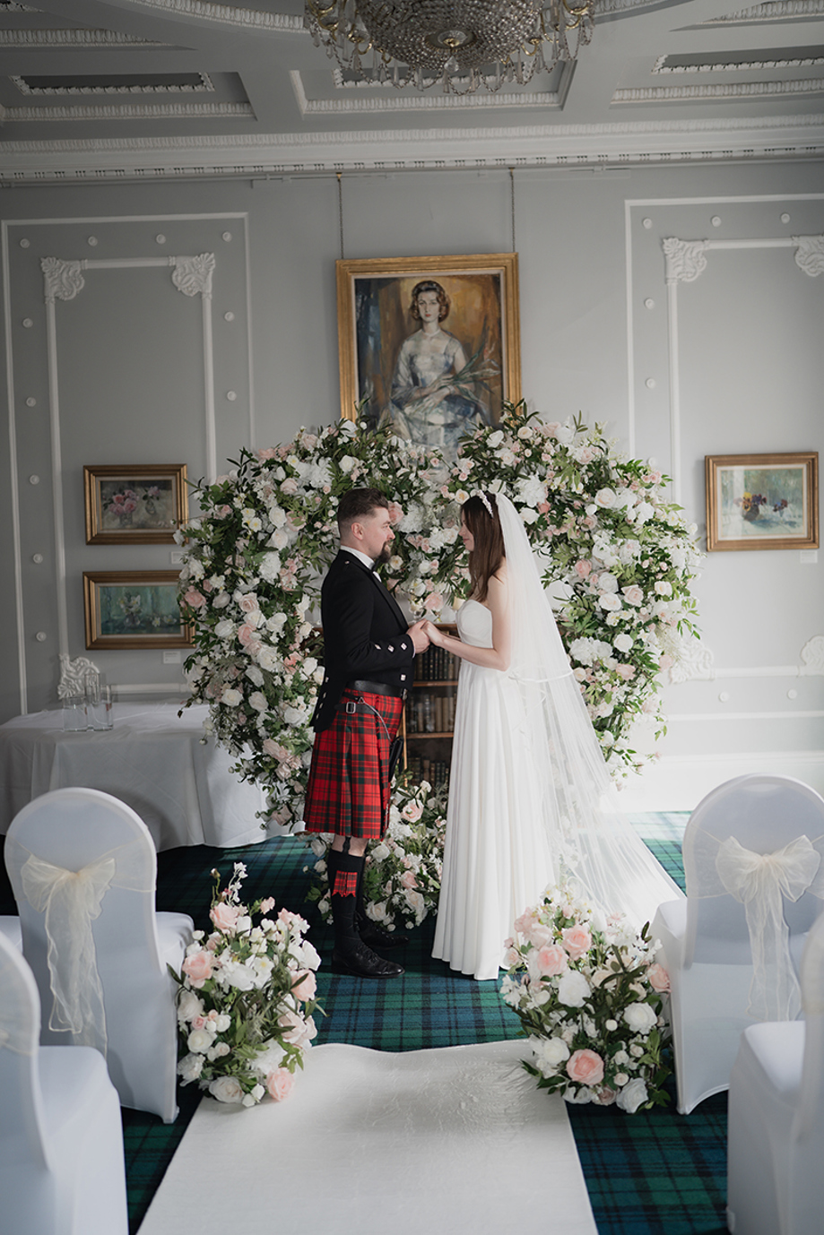 Bride and groom at altar, holding hands in front of a love heart shaped flower arch.