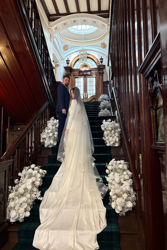 Bride and groom look back as they walk up flower-lined stairs with train behind.