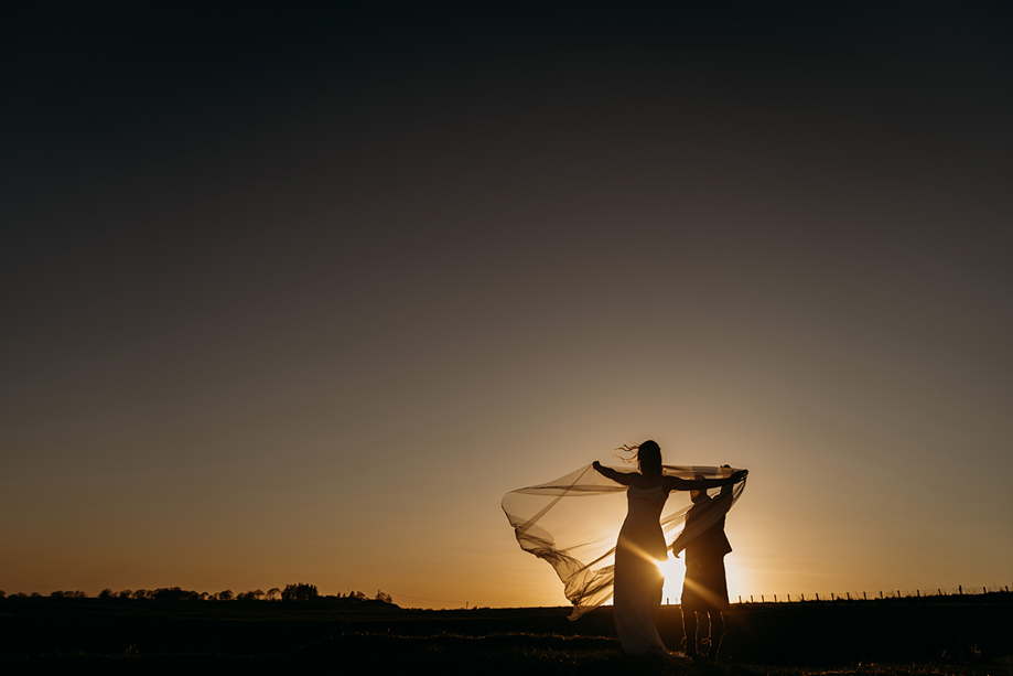 Far away image of groom and bride's shadows as brides veil blows in wind at sunset.