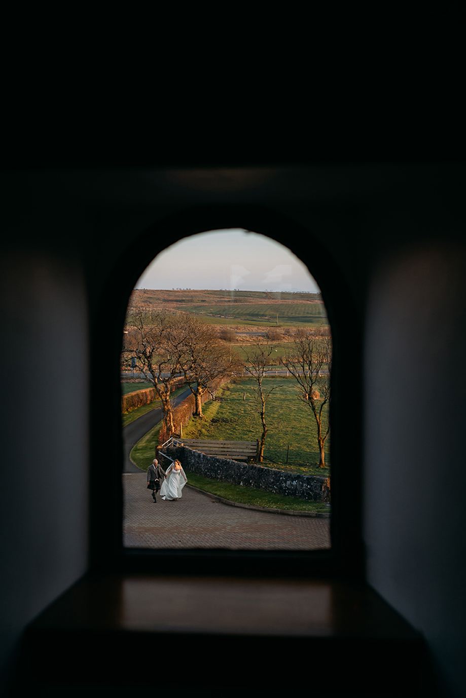 Image through arched window of bride and groom walking up driveway.