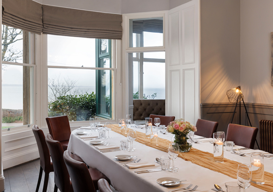 Wedding meal set up in the Macallan Room at Hotel du Vin St Andrews in Fife. Bay windows show the stunning views and the table is decorated with a mustard table runner and a small bouquet in the centre.