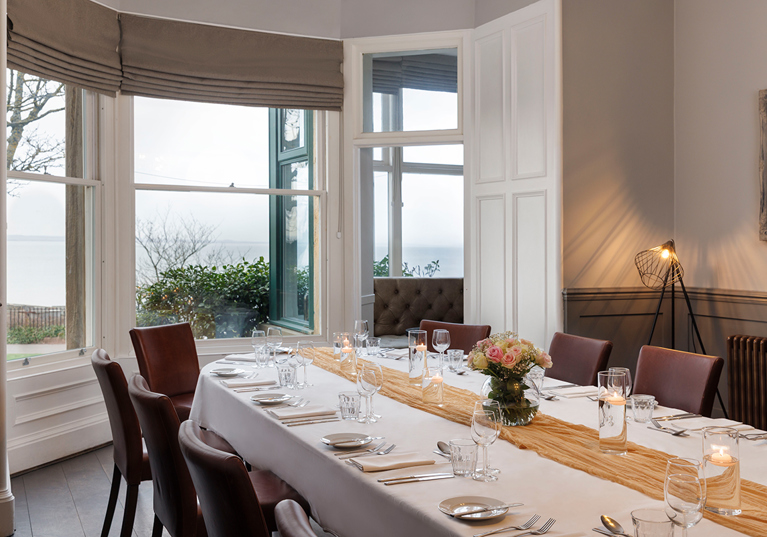 Wedding meal set up in the Macallan Room at Hotel du Vin St Andrews in Fife. Bay windows show the stunning views and the table is decorated with a mustard table runner and a small bouquet in the centre.