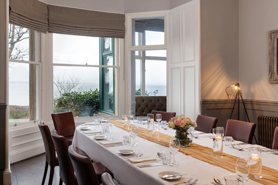 Wedding meal set up in the Macallan Room at Hotel du Vin St Andrews in Fife. Bay windows show the stunning views and the table is decorated with a mustard table runner and a small bouquet in the centre.