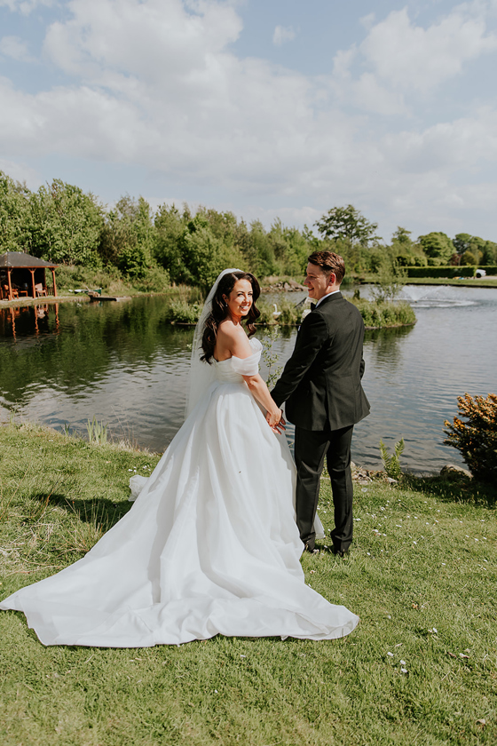 Newlywed couple holding hands beside loch at Ingliston Estate and Country Club wedding venue