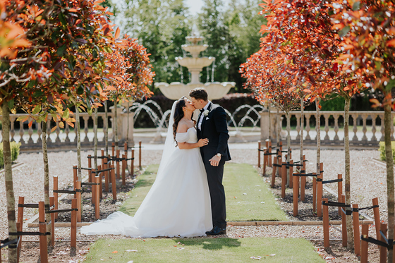 Newlywed couple kissing beside fountain and landscaped gardens at Ingliston Estate and Country Club wedding venue
