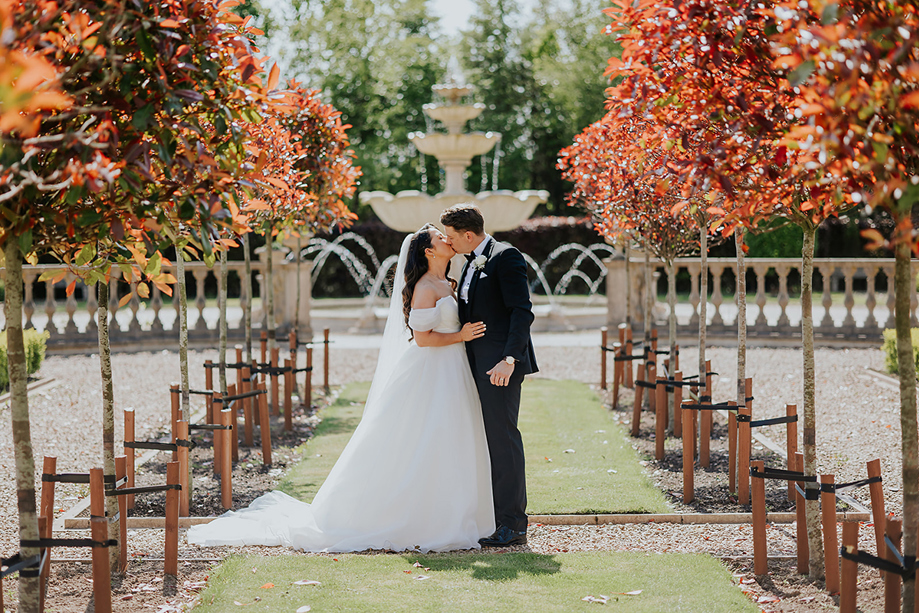 Newlywed couple kissing beside fountain and landscaped gardens at Ingliston Estate and Country Club wedding venue