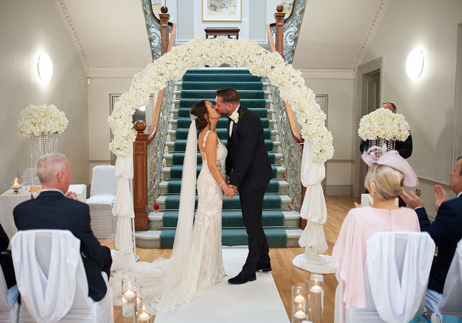 Bride and groom kiss under floral arch at altar with staircase in background