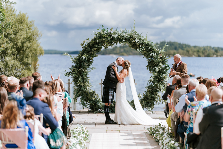 Bride and groom kiss at altar in waterside wedding ceremony with green hoop arch in background.
