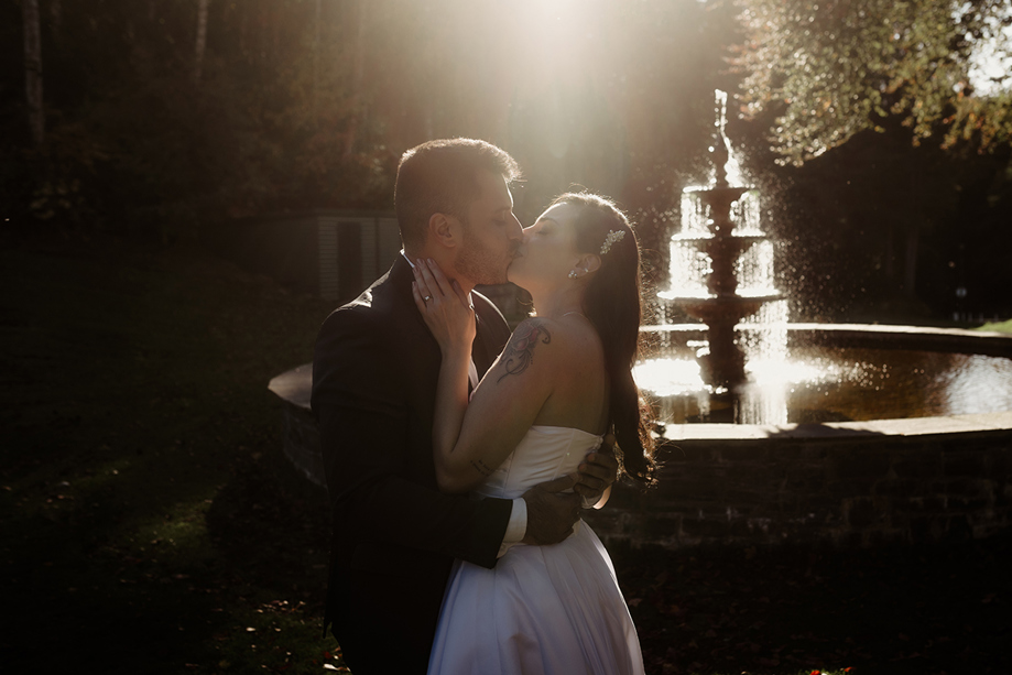 Bride and groom kissing in front of a sunlit outdoor fountain during their wedding photoshoot at Atholl Palace Hotel in Pitlochry.