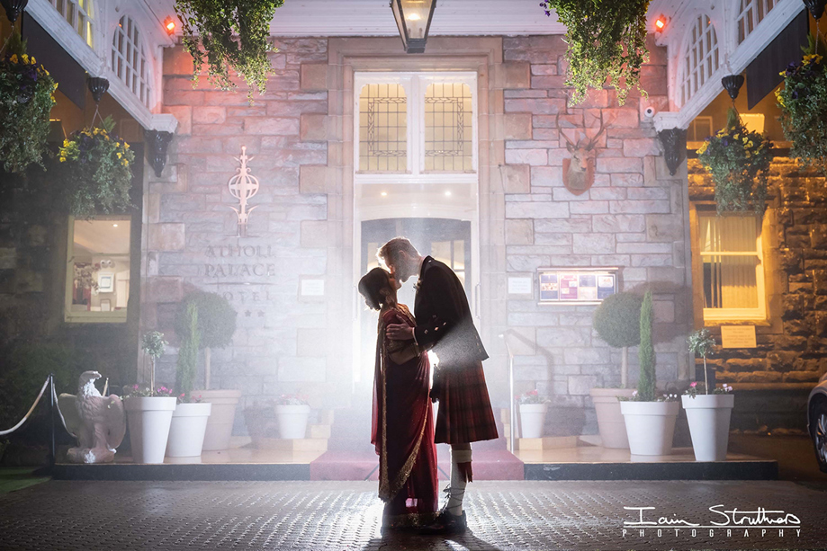 Bride and groom share a dramatic nighttime kiss outside Atholl Palace Hotel, backlit by soft mist and venue lighting at the historic stone entrance.