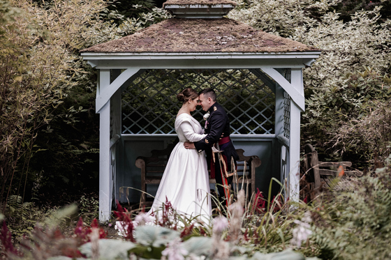 Bride and groom sharing an intimate moment beneath a rustic wooden gazebo in the gardens of Atholl Palace Hotel, surrounded by lush foliage and flowers.