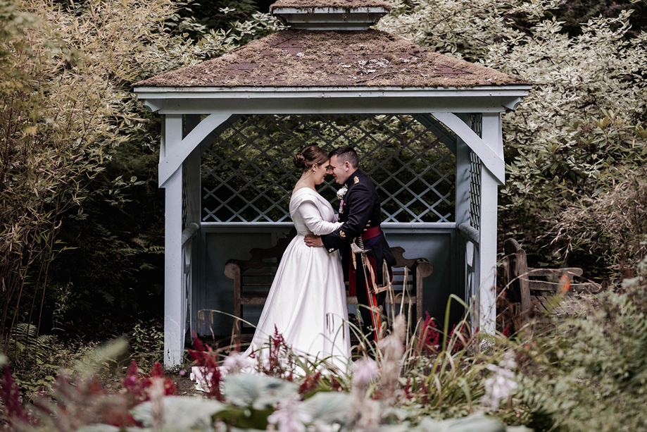 Bride and groom sharing an intimate moment beneath a rustic wooden gazebo in the gardens of Atholl Palace Hotel, surrounded by lush foliage and flowers.