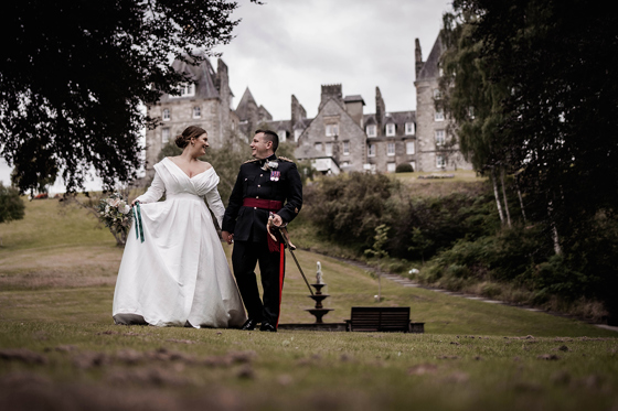 Bride and groom walking hand in hand in the gardens of Atholl Palace Hotel, with the historic Scottish castle-style hotel in the background; bride in a classic off-the-shoulder gown and groom in military dress uniform.