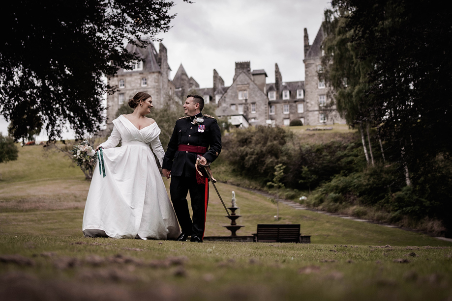 Bride and groom walking hand in hand in the gardens of Atholl Palace Hotel, with the historic Scottish castle-style hotel in the background; bride in a classic off-the-shoulder gown and groom in military dress uniform.