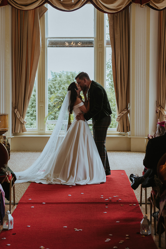 Bride and groom share their first kiss during an indoor wedding ceremony at Atholl Palace Hotel, standing on a red aisle runner in front of large windows with natural light.