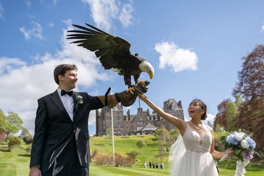 Bride and groom at Atholl Palace Hotel enjoying a falconry moment, with the groom holding a bald eagle on a glove and the bride reaching out while smiling, set against the hotel’s gardens and historic Scottish backdrop.