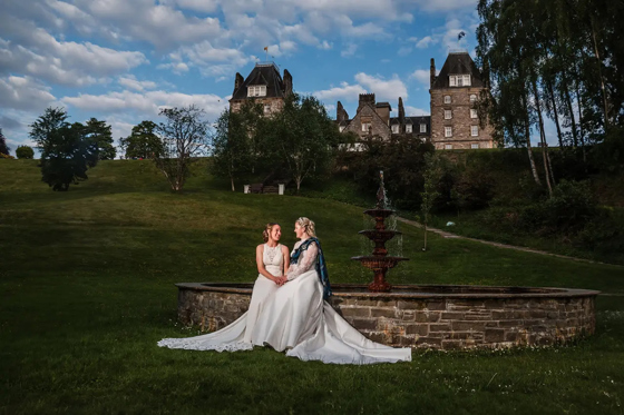 Two brides in elegant wedding dresses sit by the grand fountain at Atholl Palace Hotel, with landscaped grounds in the background.