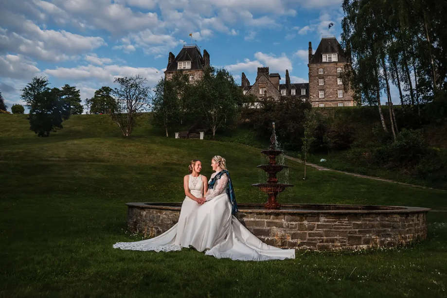 Two brides in elegant wedding dresses sit by the grand fountain at Atholl Palace Hotel, with landscaped grounds in the background.