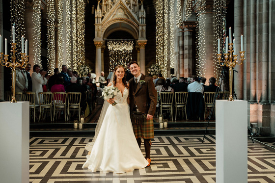 Bride and groom smiling with guests and fairy lights in background, with golden candelabras on stands in foreground