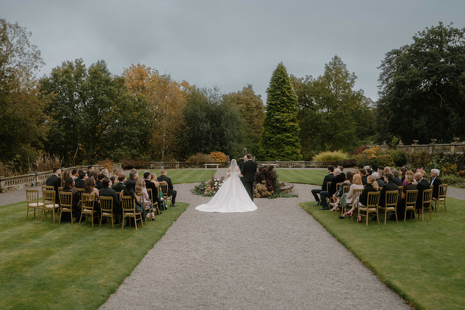 Outdoor wedding ceremony in the gardens at Auchen Castle with guests seated on gold chairs.
