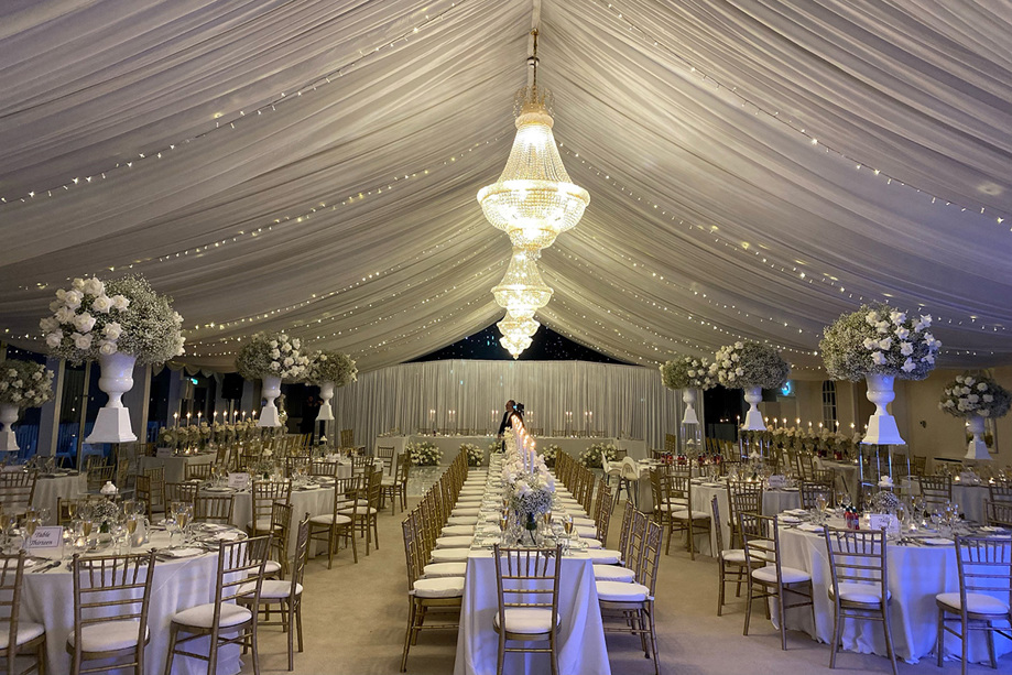 Long tables and round tables set up for wedding meal with chandeliers and draped ceiling with fairy lights