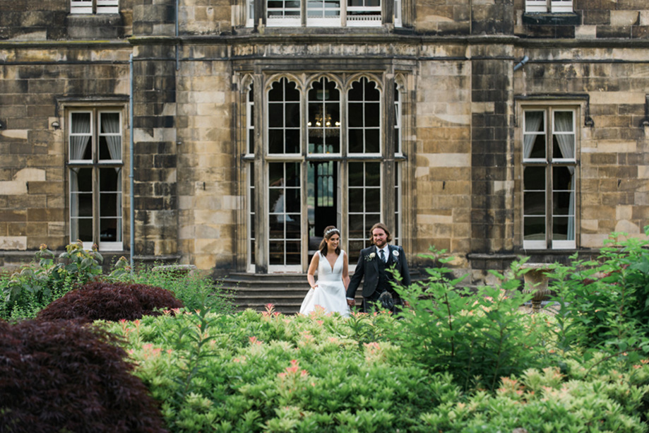 Bride and groom leaving building into gardens smiling and holding hands 