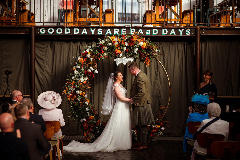 Bride and groom stand in front of large floral hoop at BAaD in Glasgow, with "Good Days Are BAaD Days" on wall in background.
