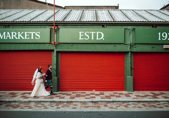 Bride and groom walk through Barra's Market in Glasgow, with red shutters behind them and "Markets Estd 1921" above them.