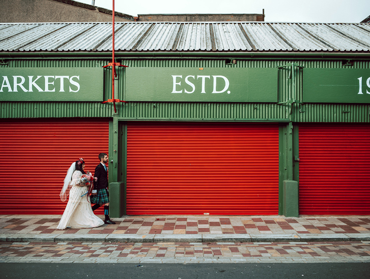 Bride and groom walk through Barra's Market in Glasgow, with red shutters behind them and "Markets Estd 1921" above them.