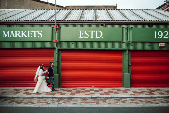 Bride and groom walk through Barra's Market in Glasgow, with red shutters behind them and "Markets Estd 1921" above them.