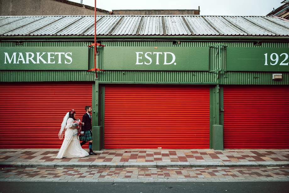 Bride and groom walk through Barra's Market in Glasgow, with red shutters behind them and "Markets Estd 1921" above them.