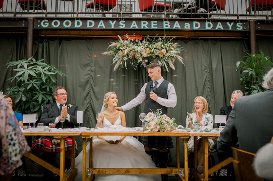 Groom's speech at top table with hand on his bride's shoulder and "Good Days Are BAaD Days" on wall behind them with flower decor.