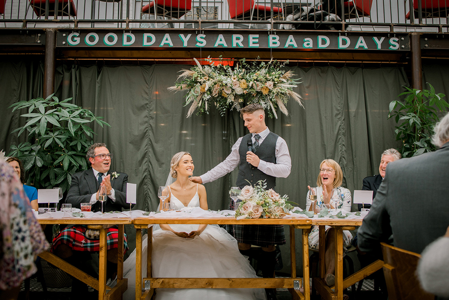 Groom's speech at top table with hand on his bride's shoulder and "Good Days Are BAaD Days" on wall behind them with flower decor.