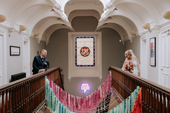 Bride and groom sharing a moment on opposite sides of the grand staircase at Netherbyres House, framed by stained glass