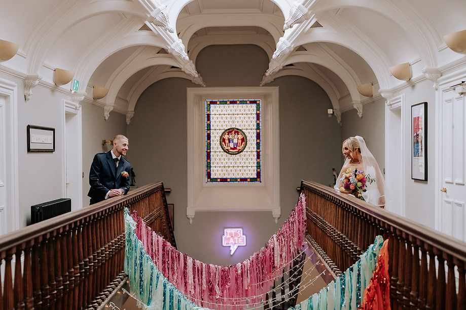 Bride and groom sharing a moment on opposite sides of the grand staircase at Netherbyres House, framed by stained glass